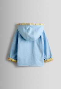 Gaiši zils kapucveida jaka ar baltām punktiņām, dzeltenas ziedu akcentētas aproces un volāna dzeltena oderējuma odere apkārt kapucim.