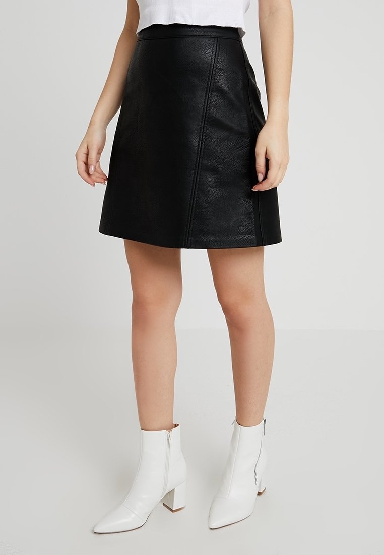 Femme portant une jupe en cuir noir, un haut blanc sans manches et des bottines blanches, debout devant un fond clair uni.