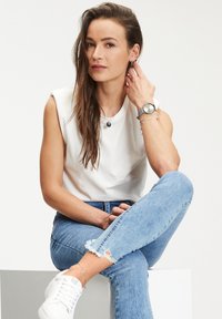 Weißes ärmelloses Oberteil, blaue verwaschene Jeans, weiße Sneaker. Silberschmuck umfasst eine Uhr und Halsketten. Haare offen mit einem natürlichen Look.