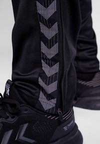 Zwarte trainingsbroek met een zijrits, voorzien van een reflecterend zigzagpatroon. Het materiaal is glad en rekbaar, gecombineerd met zwarte sneakers.