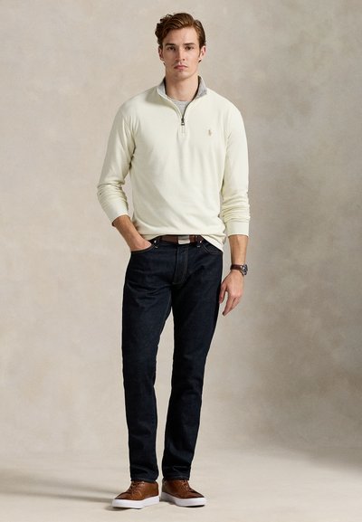 Tânăr stând, purtând un pulover crem cu fermoar până la un sfert, blugi închiși la culoare, curea maro, adidași maro asortați și ceas la încheietura mâinii stângi.