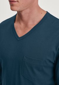 Chemise à manches longues en coton bleu foncé à col en V ; elle présente une poche sur la poitrine gauche et une texture lisse. Coutures visibles le long des bords.