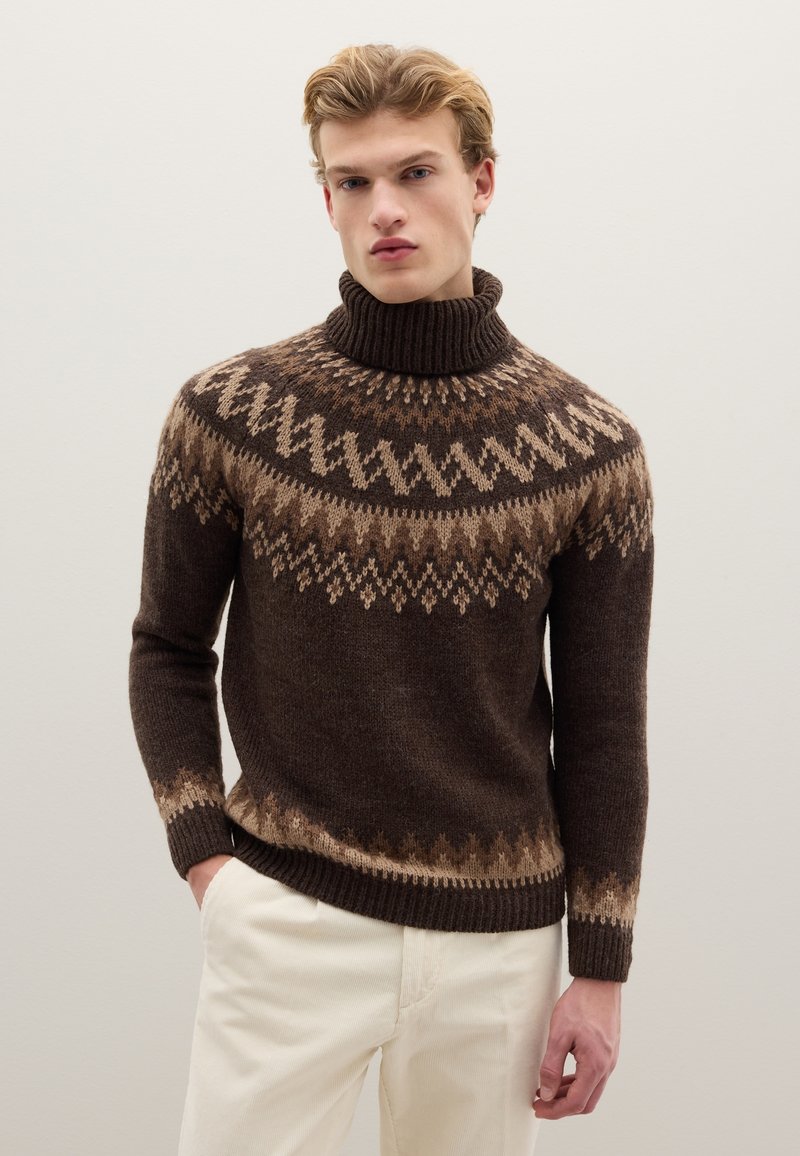 Brown turtleneck sweater with a textured knit, featuring geometric patterns in light brown. Ribbed cuffs and hem. Paired with light-colored pants.