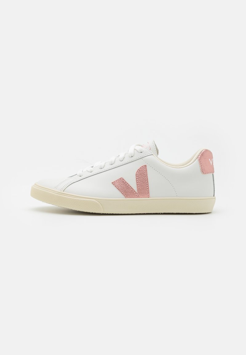 Valkoiset nahkaiset lenkkarit, joissa on teksturoitu vaaleanpunainen "V" -logo ja kantatappi, beige kumipohja, litteät nauhat ja minimalistinen muotoilu.