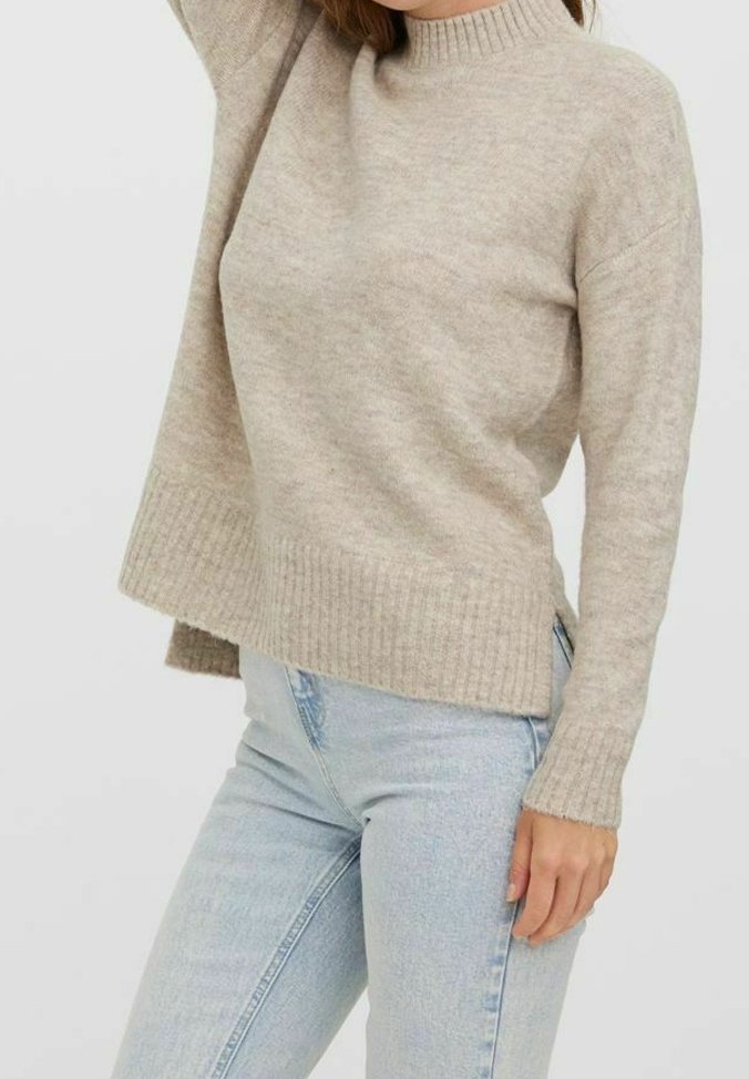 Femme portant un pull en maille beige avec des poignets et un ourlet côtelés, associé à un jean taille haute bleu clair, sur un fond uni.