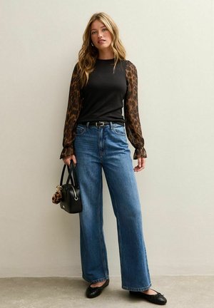 Zwart top met doorschijnende leopardenmouwen, gecombineerd met wijde blauwe jeans. Accessoires zijn een zwarte handtas en zwarte platte schoenen.