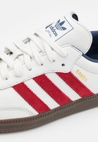 Witte sneakers met rode suède strepen, textielleer, een gemarkeerde tong en een bruine rubberen zool; heeft het woord "SAMBA" in goud. 