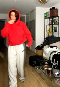 Person i rød hettegenser og hvite joggebukser går i et rotete stue med glass sofabord, bokhylle og sofa med klær.
