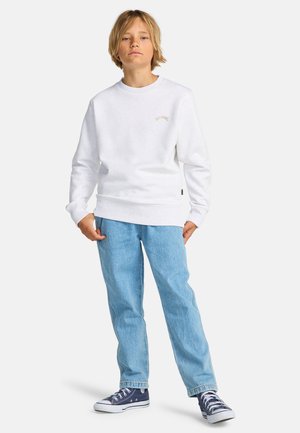 Sweatshirt blanc à col rond et poignets côtelés, associé à un jean en denim bleu clair. Des baskets noires et blanches complètent la tenue.