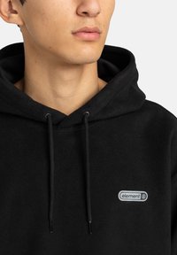 Felpa nera in pile con coulisse, caratterizzata da una piccola etichetta grigia sul petto. Tessuto morbido e vestibilità rilassata, design minimalista.