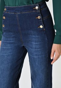 Jean taille haute bleu foncé avec des boutons dorés à l'avant, couture visible et texture en denim lisse.