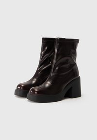 Botas de tobillo brillantes de color marrón oscuro con gruesos tacones de plataforma negros y punteras redondeadas, sobre un fondo claro y liso.