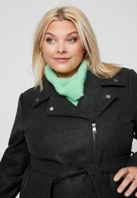 Blondinka nosi srebrne obroče za ušesa, mint zelenu pletenu turtleneck majicu, tamnosivi kaput s pojasom, zatvaračem i dugmadi, gleda desno.