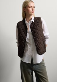 Gilet marrone trapuntato realizzato in materiale morbido, con bottoni a pressione e una vestibilità rilassata, indossato sopra una camicia bianca e abbinato a jeans blu.