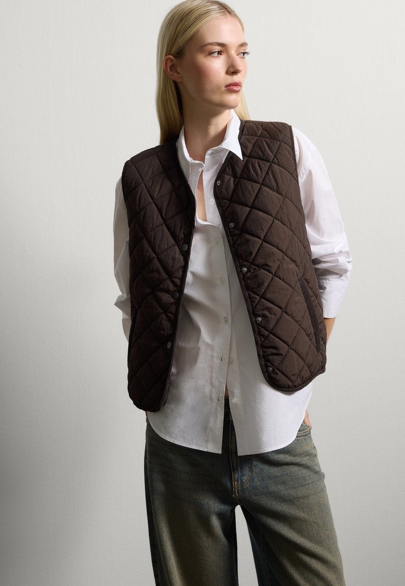 Gilet marrone trapuntato realizzato in materiale morbido, con bottoni a pressione e una vestibilità rilassata, indossato sopra una camicia bianca e abbinato a jeans blu.