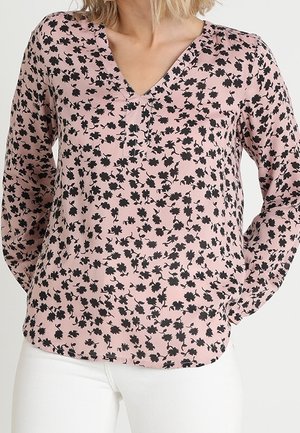 Nainen pukeutunut vaaleanpunaiseen, pitkähihainen V-kaula-aukolliseen paitaan, jossa on musta kukkakuosi, ja valkoiset housut, kädet lepäävät selän takana.