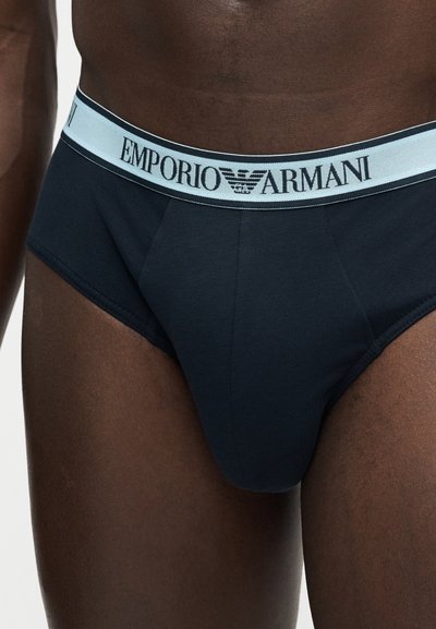 Ανδρικός κορμός που φοράει μαύρα εσώρουχα με ανοιχτό μπλε ζωνάρι μέσης, στο οποίο βρίσκεται το λογότυπο Emporio Armani και το έμβλημα αετού.