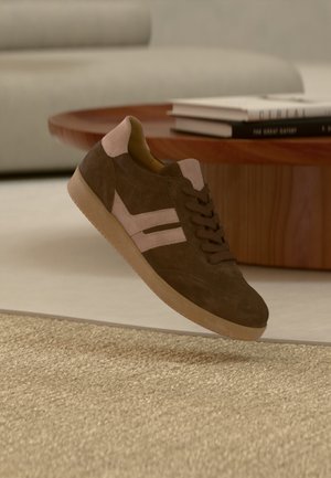 Baskets en daim marron avec des accents beiges flottant au-dessus d'un tapis beige texturé, avec une table basse en bois et des livres en arrière-plan.