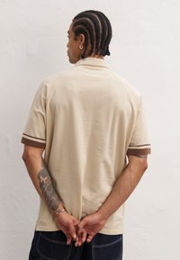 Beige poloshirt med korte raglanærmer, som har brune stribede accenter. Blød tekstur, afslappet pasform og en subtil detalje med en rygsøm.