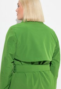 Trench coat impermeabile verde con vita regolabile e dettaglio posteriore a strati. Texture liscia e design su misura.
