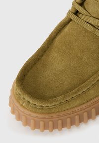 Chaussure en suede vert avec un bout arrondi, des détails cousus et une semelle en caoutchouc tan épaisse et texturée. Les lacets sont visibles sur le dessus.