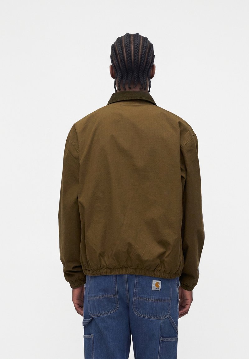 Homme aux cheveux tressés portant une veste vert olive et un jean bleu, debout, le dos tourné, devant un fond clair uni.