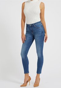 Mujer luciendo una blusa sin mangas blanca con textura, jeans ajustados azules, tacones desnudos y una delicada pulsera de oro en su muñeca izquierda.