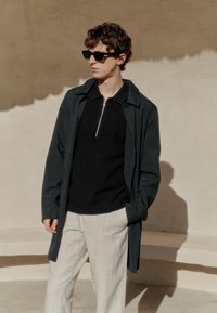 Mladenič, ki nosi črna sončna očala, temno modro jakno, črn pulover z zadrgo in bele hlače, stoji ob bež steni na sončni svetlobi.