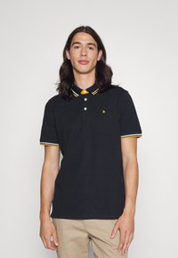 Camisa polo preta feita de algodão com uma pala de dois botões, colarinho listrado em amarelo e branco e bordas das mangas, apresentando um pequeno logotipo no peito.