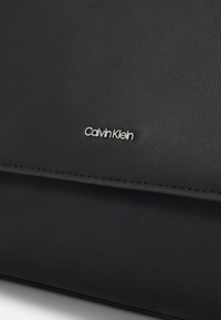 Bolso de piel negro con una textura suave y costuras mínimas. Presenta un logo plateado de "Calvin Klein" en la parte frontal.