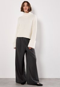 Maglione bianco a collo alto lavorato a maglia con texture a coste, abbinato a pantaloni larghi grigi scuri a righe. Ballerine nere completano l'outfit.