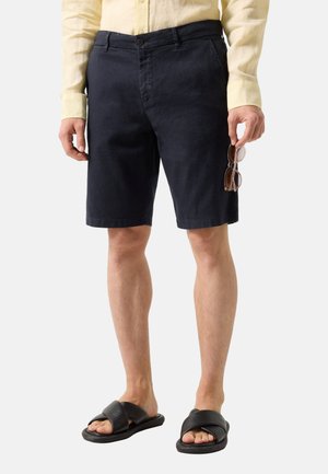 Mand iført sorte knælange shorts, gul langærmet skjorte, sorte slidesandaler, holder solbriller i højre hånd.