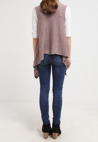 Langer, ärmelloser Cardigan aus leichtem mauvefarbenem Strickstoff, der hinten drapiert ist und offene Seiten hat, kombiniert mit blauen Skinny-Jeans und schwarzen High-Heels-Stiefeln.