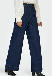 Pantalons en denim larges bleu foncé avec des coutures visibles et des poches, associés à un chemisier crème et des bottes à talons noires.