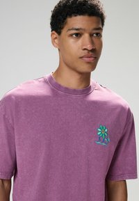 Lila Übergröße T-Shirt aus weichem Baumwollmaterial, mit einem bunten gestickten Blumendesign auf der linken Brust und einem Rundhalsausschnitt.