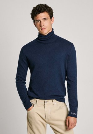 Suéter de cuello alto azul marino hecho de tela suave, con mangas largas y textura acanalada, con el logo "PEPE" en el puño.