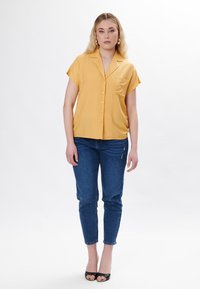 Camicia a maniche corte con bottoni in tessuto giallo brillante, dotata di taschino sul petto, abbinata a jeans slim-fit blu scuro e sandali neri con apertura sulla punta.