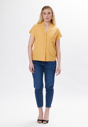 Camicia a maniche corte con bottoni in tessuto giallo brillante, dotata di taschino sul petto, abbinata a jeans slim-fit blu scuro e sandali neri con apertura sulla punta.