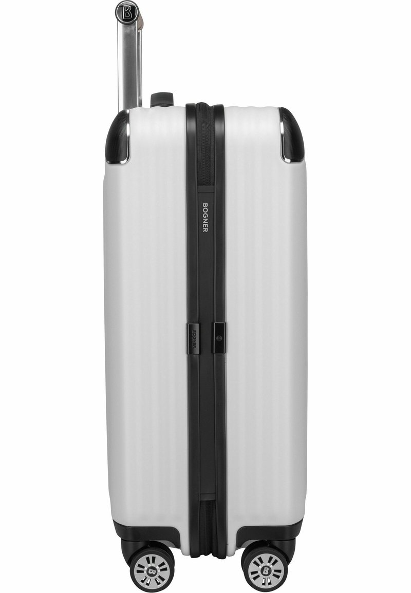 Biała, twarda walizka z czarnymi akcentami, pionowym rowkowanym wzorem, podwójnymi zamkami błyskawicznymi oraz czterema obrotowymi kółkami, co ułatwia transport.