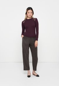 Langärmliges burgunderfarbene Oberteil mit Spitzenakzenten an den Schultern, kombiniert mit weit geschnittenen grauen Hosen. Die Schuhe haben ein zweifarbiges Design.