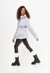 Giacca imbottita grigio chiaro con vita elasticizzata, dotata di cappuccio e tasche frontali. Abbinata a leggings neri e stivali neri massicci.
