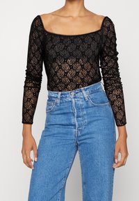 Schwarzes Langarmoberteil aus Spitze mit floralem Muster, quadratischem Ausschnitt und tailliertem Schnitt, kombiniert mit hochgeschnittenen blauen Jeans.