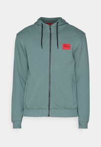 Mikina s kapucí v tyrkysové barvě s celým zipem, s červenou čtvercovou nášivkou loga, černými šňůrkami a texturovanou tkaninou.