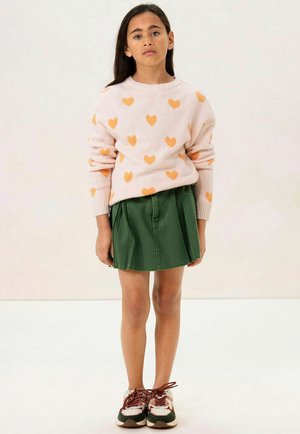 Pull léger rose avec des motifs de cœurs orange, associé à une jupe plissée verte. Les chaussures présentent des accents marron et blanc avec des lacets.