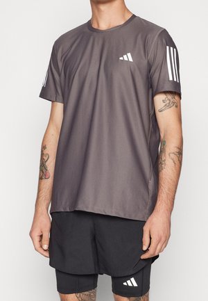 Mann trägt dunkelgraues Adidas kurzärmeliges Shirt mit weißen Streifen auf den Ärmeln und schwarzen Shorts, zeigt Tattoos auf beiden Armen.