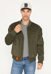 Veste bomber en velours côtelé vert olive avec des poignets côtelés, fermeture éclair à l'avant et poches latérales ; détail d'étiquette orange en bas à gauche. T-shirt gris en dessous.