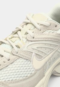 Αθλητικό παπούτσι ανοιχτού μπεζ χρώματος με υλικά από δίχτυ και σουέτ, διακριτή υφή και καμπυλωτό σχέδιο με χαρακτηριστικό λογότυπο στη πλευρά.