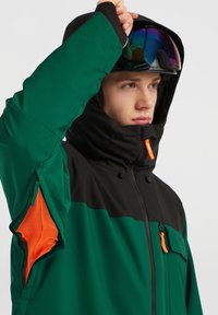 Grøn og sort jakke med hætte, der har orange accenter, netunderarmsventilation og en lynlåslomme på fronten.