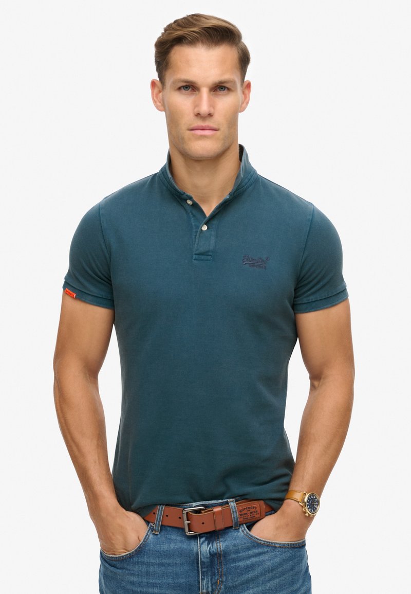 Camisa polo de algodón en color verde azulado con una tapeta de botones, mangas cortas y un logo discreto en el lado izquierdo del pecho. Lleva un cinturón marrón y jeans de mezclilla.