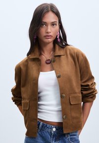 Veste en suede marron avec un col, fermeture à boutons et deux poches avant. Assortie avec un débardeur blanc et un jean en denim.